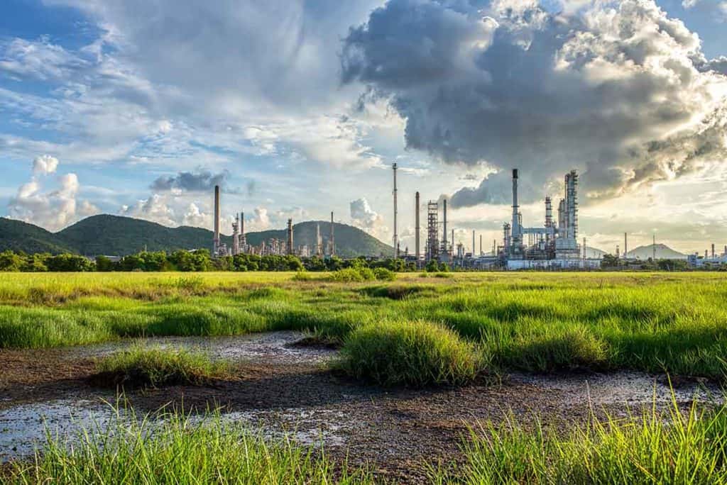 A lush green field with patches of water in the foreground and an industrial refinery complex with tall chimneys in the background, set against hills and a dramatic sky with large clouds.