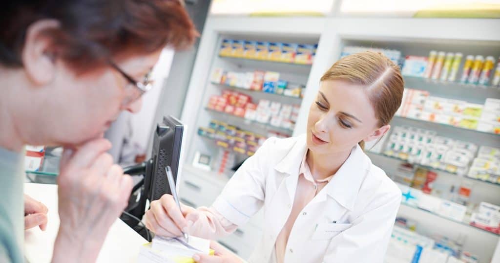A pharmacist in a white coat assists a customer at the counter, writing on a notepad. Shelves behind them are stocked with various medications and health products.