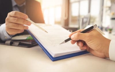 Two people sitting at a desk review and discuss a document on a clipboard. One person points with a pen while the other holds a pen, ready to write or sign. The setting is a bright office space.