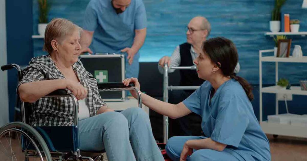 A nurse kneels beside an elderly woman in a wheelchair, offering comfort. In the background, another nurse attends to a senior man with a walker. Medical supplies and shelves are visible in the room.