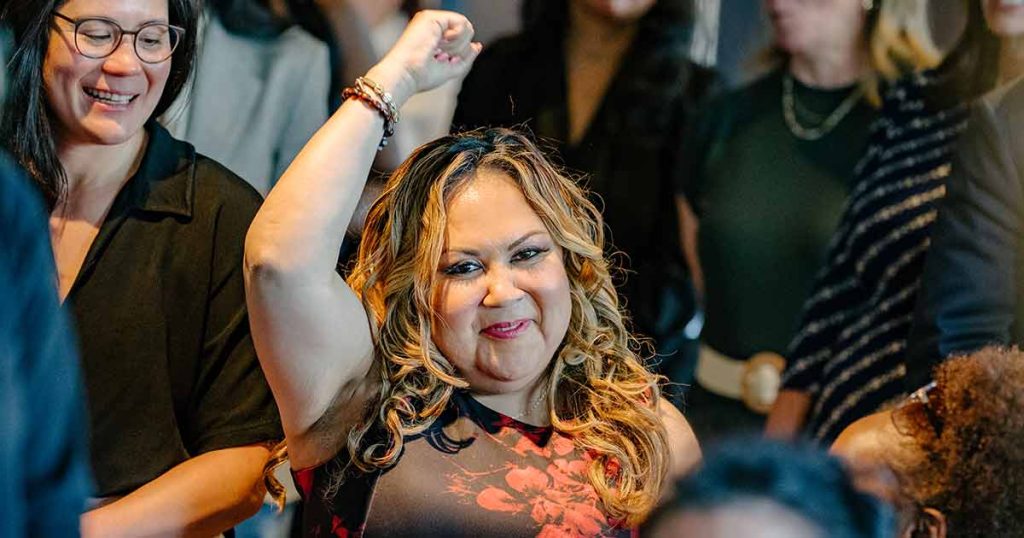 A smiling woman with long, wavy hair raises her fist in celebration or victory among a group of people, creating a joyful and empowering atmosphere.