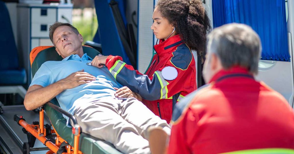 A paramedic checks the pulse of a conscious man lying on a stretcher inside an ambulance, while another paramedic sits nearby with his back to the camera.