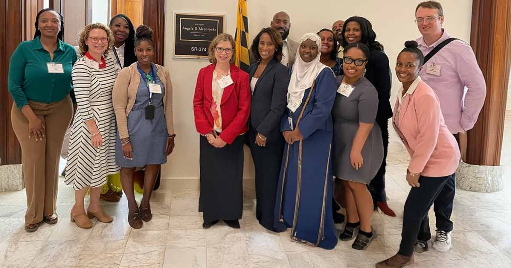 A diverse group of thirteen people, dressed in business and professional attire, smiling and posing together indoors in front of an office door with a nameplate and flag.