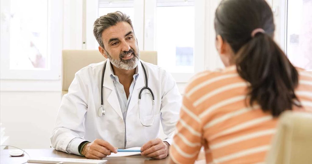 A male doctor with a stethoscope around his neck sits at a desk, smiling and talking to a patient wearing a striped shirt, in a bright medical office.