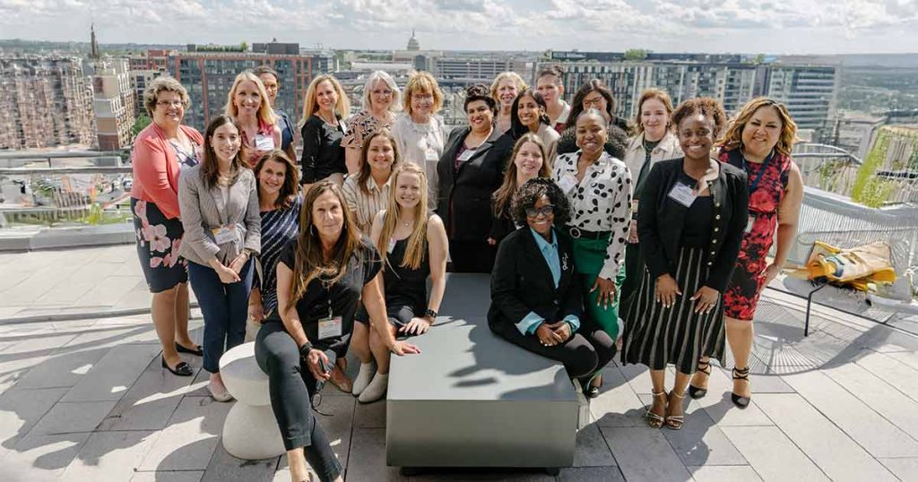 A group of around 25 women, dressed in business attire, pose and smile together on a rooftop patio with a cityscape and blue sky in the background.