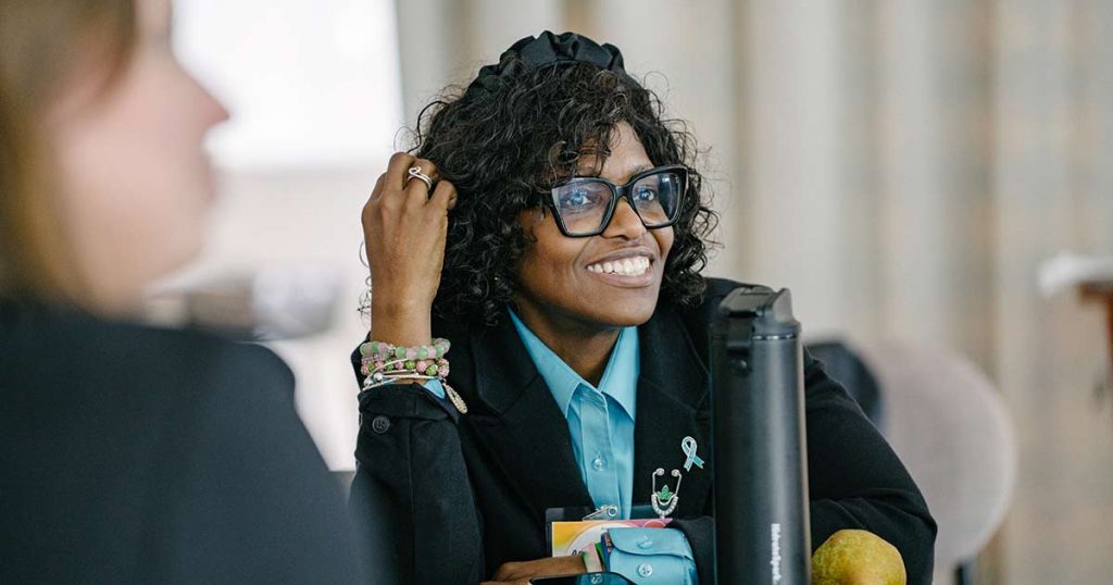 A woman with curly hair and glasses, wearing a blue shirt and black blazer, smiles while sitting at a table. She has beaded bracelets on her wrist and a ribbon pin on her jacket. The background is blurred.