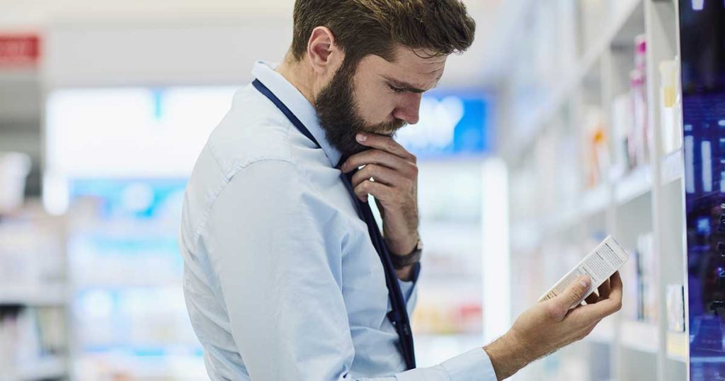 Man dressed in business attire is in a pharmacy aisle and looking intently at a drug package label.