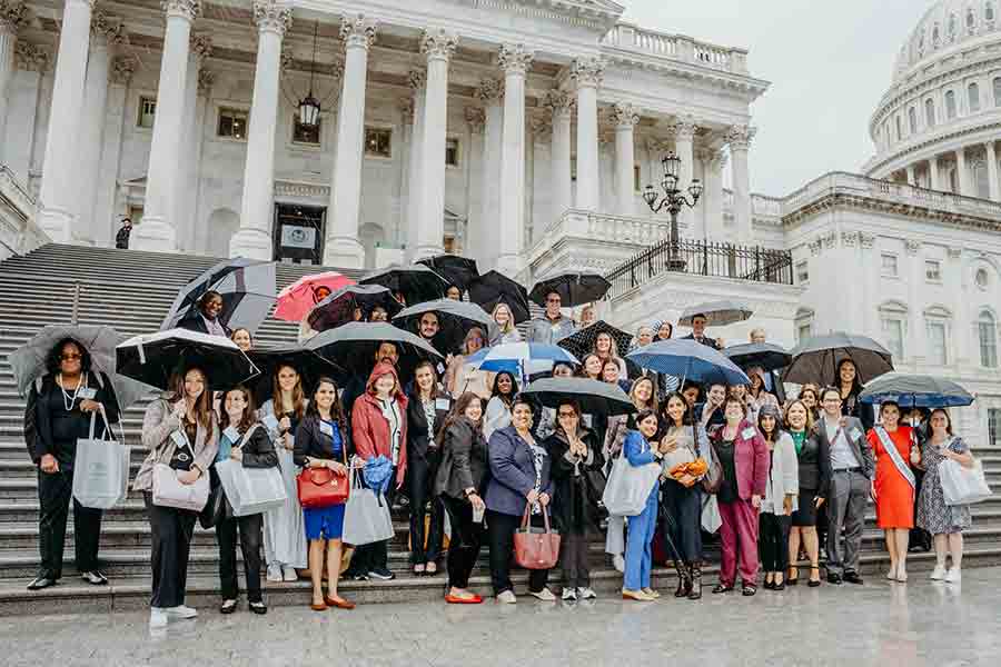 A large group of advocate stand on the steps of a government building, holding umbrellas on a rainy day. The group appears diverse and is posing together, with the U.S. Capitol building visible in the background.
